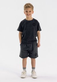 Mörkgrå bomullströja med korta ärmar och en subtil logotyp; matchande shorts med fickor. Vita sneakers och randiga ankelstrumpor.