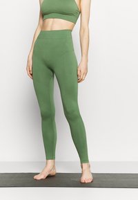 Woman in green seamless high-waisted leggings and matching sports bra standing barefoot on a yoga mat against a white wall.