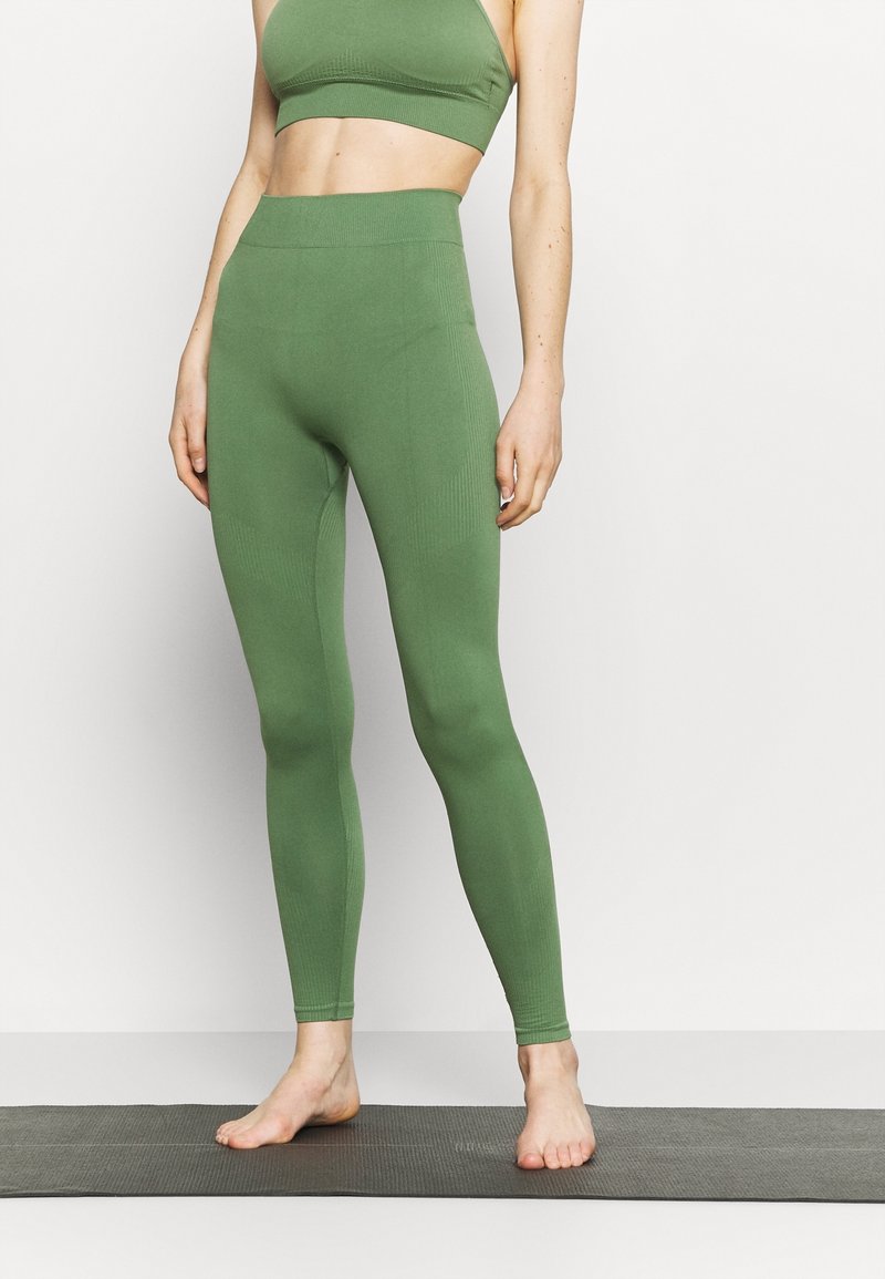Woman in green seamless high-waisted leggings and matching sports bra standing barefoot on a yoga mat against a white wall.