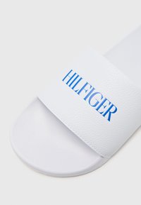 Bijele kožne sandale s potplatom na klizanje, s teksturiranim trakama koje sadrže utisnuti plavi logo "HILFIGER", zaobljenim potplatom i minimalističkim dizajnom.