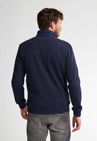 Felpa blu navy con colletto alto, polsini e vita a coste. Texture liscia con vestibilità ampia, indossata sopra pantaloni grigi.