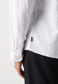 Camisa blanca de lino con puños abotonados, presenta una etiqueta lateral y un corte relajado, mostrando un tejido texturizado y arrugas sutiles.
