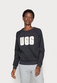 Crna dukserica s velikim UGG logotipom u krem boji, raglan rukavi i rebrasti manžeti. Uklapaju se s sivim trenirkama. Glatka tekstura.