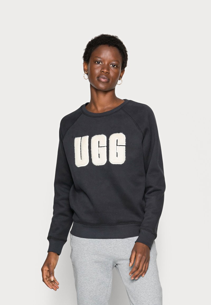 Crna dukserica s velikim UGG logotipom u krem boji, raglan rukavi i rebrasti manžeti. Uklapaju se s sivim trenirkama. Glatka tekstura.