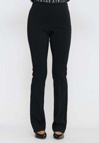 Pantaloni neri su misura indossati con scarpe nere a punta, mostrati dalla vita ai piedi su uno sfondo neutro.