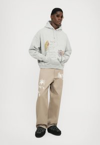 Grå sweatshirt med blomster grafik og tekst på forsiden, parret med beige bukser med hvid broderi og sorte broderede slip-on sko.