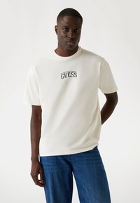 Camiseta blanca de manga corta con un logo y texto negro de "GUESS" en el pecho; cuello redondo, textura suave, combinada con unos jeans de mezclilla azul.