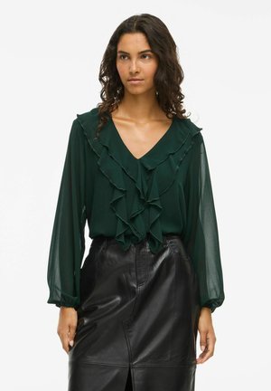 Groene blouse met een ruches ontwerp langs de halslijn en mouwen, gemaakt van lichtgewicht stof. Gecombineerd met een zwarte leren rok.