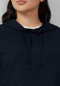 Frau in einer dunkelblauen Kapuzenjacke mit Kordeln und einem weißen Shirt, das am Ausschnitt sichtbar ist, zeigt nur Lippen und Kinn.