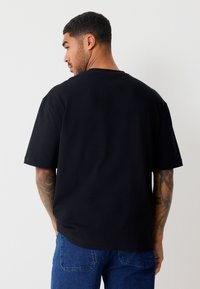 Camiseta de algodón negra, de manga corta, cuello redondo, ajuste holgado, diseño liso. El modelo lleva jeans de mezclilla azul, mostrando tatuajes en el brazo.