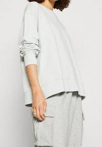 Světle šedá oversized mikina s žebrovanými manžetami a lemem, má kulatý výstřih a měkkou texturu. Nosit se s šedými cargo kalhotami.