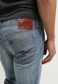 Vaqueros de mezclilla azul claro con un parche de cuero con el logo de Lee en la cintura trasera, que cuentan con un diseño clásico de cinco bolsillos y un acabado desgastado.