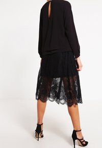 Blusa nera a maniche lunghe con apertura a chiave sul retro, abbinata a una gonna in pizzo nero trasparente. Presenta un design a strati e sandali neri con tacco alto.