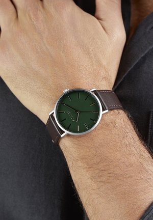 Grüne Zifferblatt-Uhr mit silbernem Gehäuse, braunem Lederarmband, minimalistische Stundenmarkierungen und einer glatten Oberfläche auf Zifferblatt und Armband.