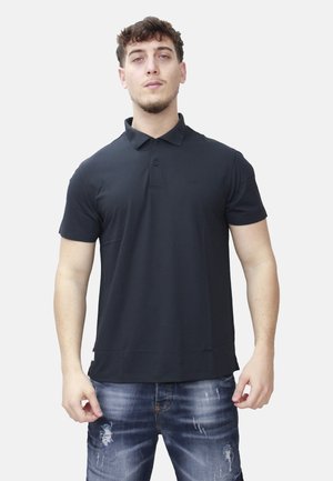 Uomo in piedi con le braccia leggermente piegate, indossa una polo nera a maniche corte e jeans blu strappati, su sfondo semplice.