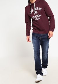 Dieprode hoodie met grijze tekst en trekkoorden, gecombineerd met donkerblauwe denim jeans. Witte sneakers maken de outfit compleet. Casual pasvorm, comfortabele stijl.