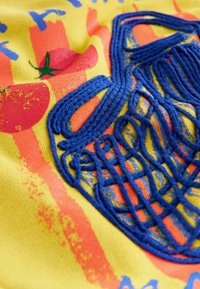 Geel stof heeft een blauw geborduurd ontwerp dat lijkt op een net, met rode tomaatafbeeldingen en oranje strepen, wat een textuureffect creëert.