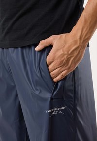 Laivastonsiniset vedenpitävät shortsit, joissa on kiiltävä pinta, sivutasku ja valkoinen "SWEDEMOUNT"-logo. Näkyvissä joustava vyötärö.