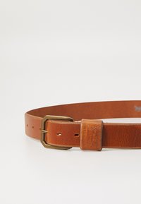 Curea din piele maro cu o cataramă din alamă. Dispune de mai multe găuri pentru ajustare și detalii cu logo gravat aproape de cataramă. Textură netedă.