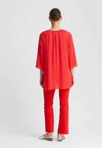 Rote, fließende Tunika mit Dreiviertelärmeln, kombiniert mit figurbetonten roten Hosen. Hausschuhe mit einem strukturierten Muster runden das Outfit ab.