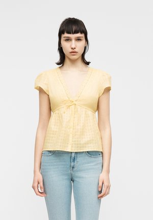 Junge Frau mit kurzen dunklen Haaren trägt eine hellgelbe Bluse mit V-Ausschnitt und Puffärmeln sowie hellblaue Jeans, sie steht vor einem weißen Hintergrund.