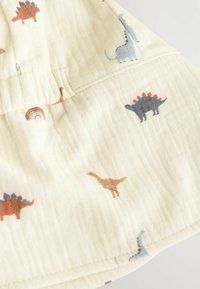 Tissu en coton de couleur jaune clair avec divers petits imprimés de dinosaures en bleu, marron et orange. Surface texturée avec des détails de couture visibles.
