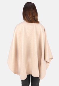 Beige Wollponcho mit einer weichen Textur, weitem Schnitt und abgerundetem Saum. Über schwarzen Leggings getragen, zeigt er ein lockeres und fließendes Design.