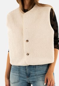 Crème fleece vest met een ronde halslijn, twee knopen aan de voorkant en een mouwloos ontwerp; gedragen over een zwarte outfit met een textuur op de oppervlakte.