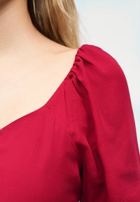 Κοντινό πλάνο στον ώμο μιας γυναίκας πο υ φοράει ένα κόκκινο ένδυμα με μανίκι πούφ με ρυτιδώσεις και φαρδιά λαιμόκοψη.