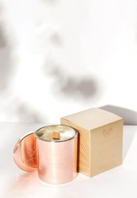 Rózowo-złoty pojemnik na świecę z drewnianym knotem, przykryty srebrną pokrywką. Towarzyszy mu jasno-brązowe pudełko z wypukłym geometrycznym wzorem.
