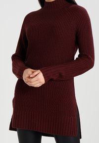 Vinröd stickad tröja med hög hals, ribbad struktur, långa ärmar och sidoflikar. Har ett geometriskt mönster på framsidan.