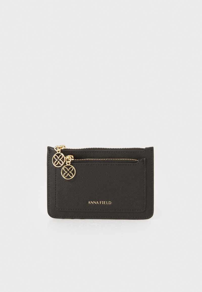 Portefeuille en synthétique noir avec une fermeture éclair dorée, deux pendentifs décoratifs et le logo en relief "ANNA FIELD" sur le devant. Design rectangulaire.