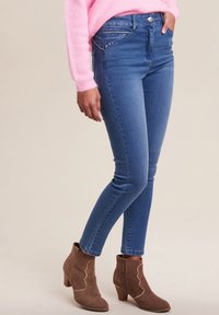 Jean skinny dans un bleu moyen avec un léger éclat, présentant des poches avant et des clous décoratifs. Assorti à des bottes cheville marron.