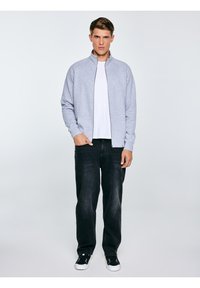 Młody mężczyzna stoi, ubrany w jasnoszarą kurtkę na zamek, białą koszulkę, luźne czarne dżinsy i czarne trampki z białymi podeszwami.