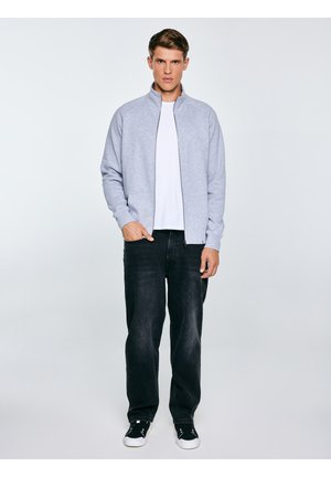 Młody mężczyzna stoi, ubrany w jasnoszarą kurtkę na zamek, białą koszulkę, luźne czarne dżinsy i czarne trampki z białymi podeszwami.