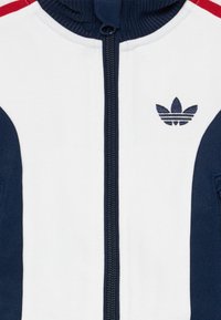 Veste de survêtement avec fermeture éclair à l'avant, col côtelé, corps blanc avec des accents navy et rouge, et logo brodé sur la poitrine.