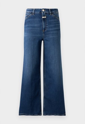 Frau steht und trägt einen cremefarbenen, geknöpften Cardigan, ein weißes Hemd mit Kragen, weit geschnittene blaue Jeans und cremefarbene High-Heels vor einfachem Hintergrund.