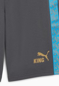 Shorts de sport gris avec une bande latérale bleue à motif, l'inscription "KING" en lettres dorées et un logo Puma doré. Tissu doux avec une texture lisse.
