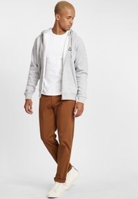 Sweat à capuche gris zippé avec un logo triangulaire, superposé à un t-shirt blanc, associé à un pantalon marron et des baskets blanches. Design simple.