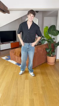Camisa preta de manga curta, jeans de denim azuis, tênis prateados. Em pé numa sala moderna com piso de madeira e uma planta verde por perto.
