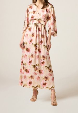 Woman wearing a light pink floral wrap dress with wide sleeves paired with beige strappy heeled sandals on a plain background.