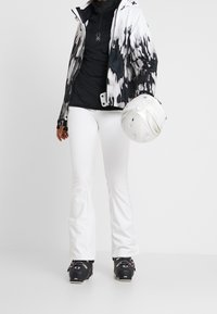 Conjunto de esquí con una chaqueta de patrón en blanco y negro, bajo negro, pantalones blancos y botas de esquí negras, sosteniendo un casco blanco.