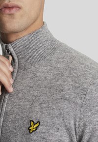 Pullover grigio con colletto alto e mezza zip. Presenta una texture a coste e un piccolo logo giallo ricamato sul petto. Materiale in maglia morbida.