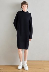 Černé pletené šaty ke kolenům s vysokým límcem a širokými rukávy, doplněné bílými teniskami. Hladká textura, jednoduchý design.