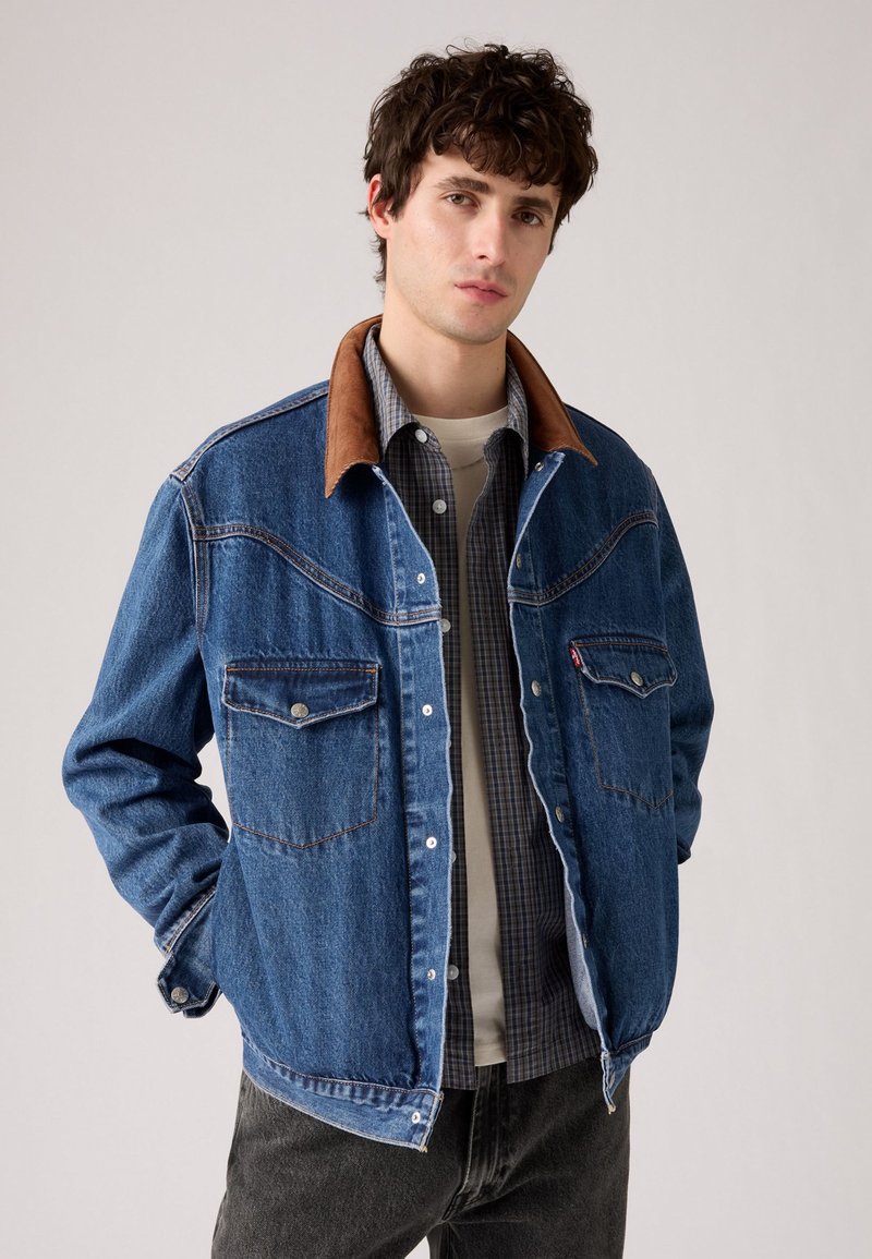 Joven con cabello rizado oscuro que lleva una chaqueta de mezclilla azul con cuello marrón sobre una camisa de cuadros y una camiseta blanca, de pie con las manos en los bolsillos.