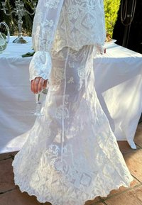 Rochie albă brodată cu mâneci lungi, cu modele florale și fustă vaporoasă. Un pahar este ținut în mâna dreaptă.