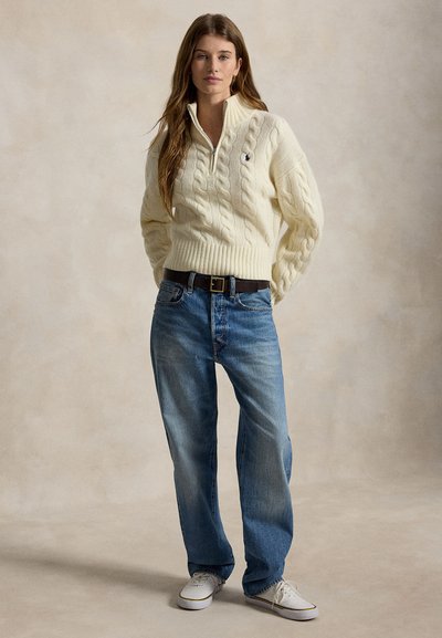Ung kvinde står, iført en cremefarvet kabelstrikket sweater med lynlås, blå jeans, sort bælte og hvide sneakers, neutral baggrund.