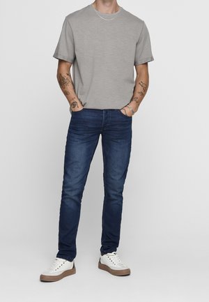 Hombre con camiseta gris clara, vaqueros ajustados azul oscuro, zapatillas blancas, con las manos en los bolsillos y tatuajes visibles en ambos antebrazos.