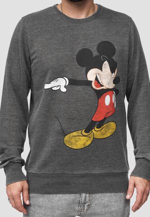 Mann trägt einen grauen Pullover mit einem großen Mickey-Mouse-Grafik im Vintage-Stil, der rote Shorts und gelbe Schuhe zeigt.
