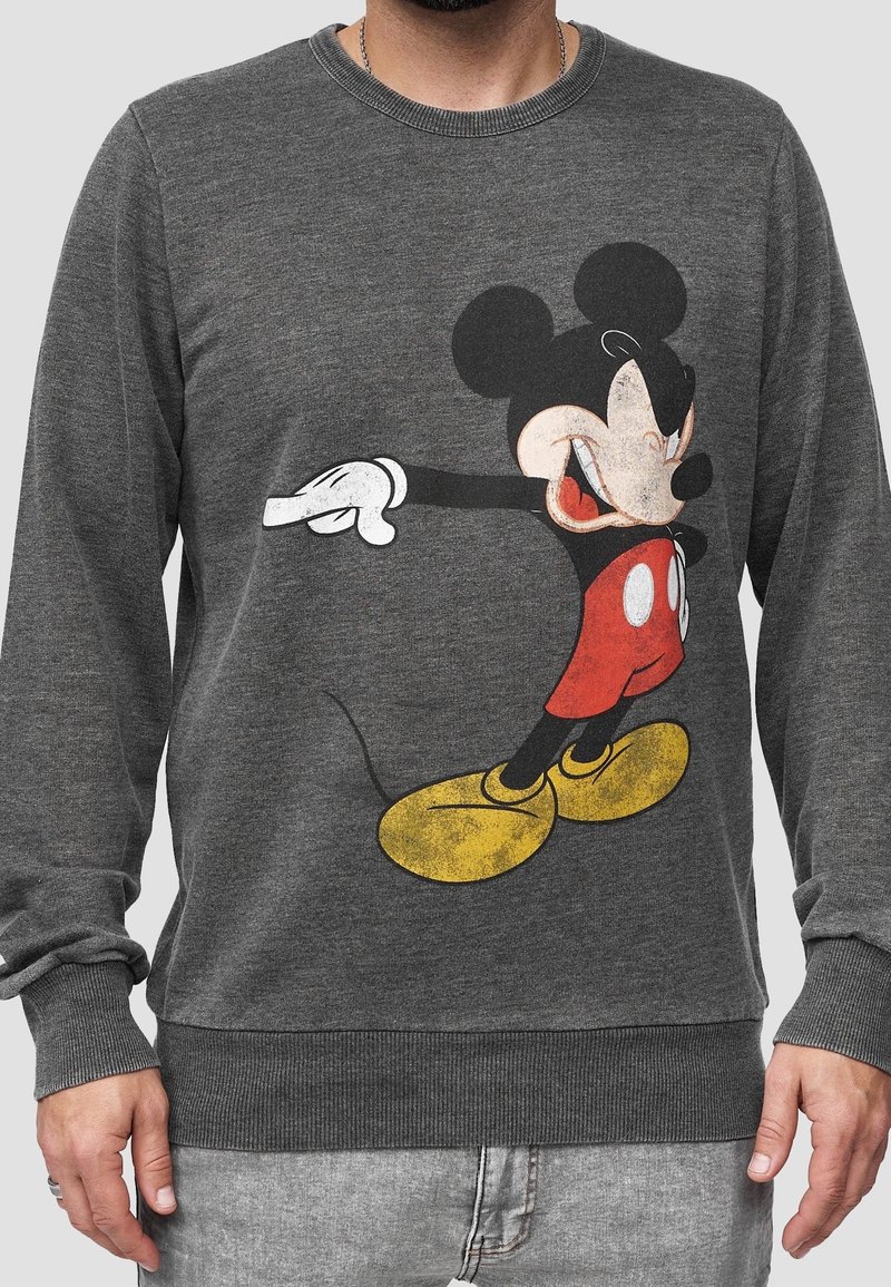 Homme portant un sweat-shirt gris avec un grand dessin vintage de Mickey Mouse en short rouge et chaussures jaunes.